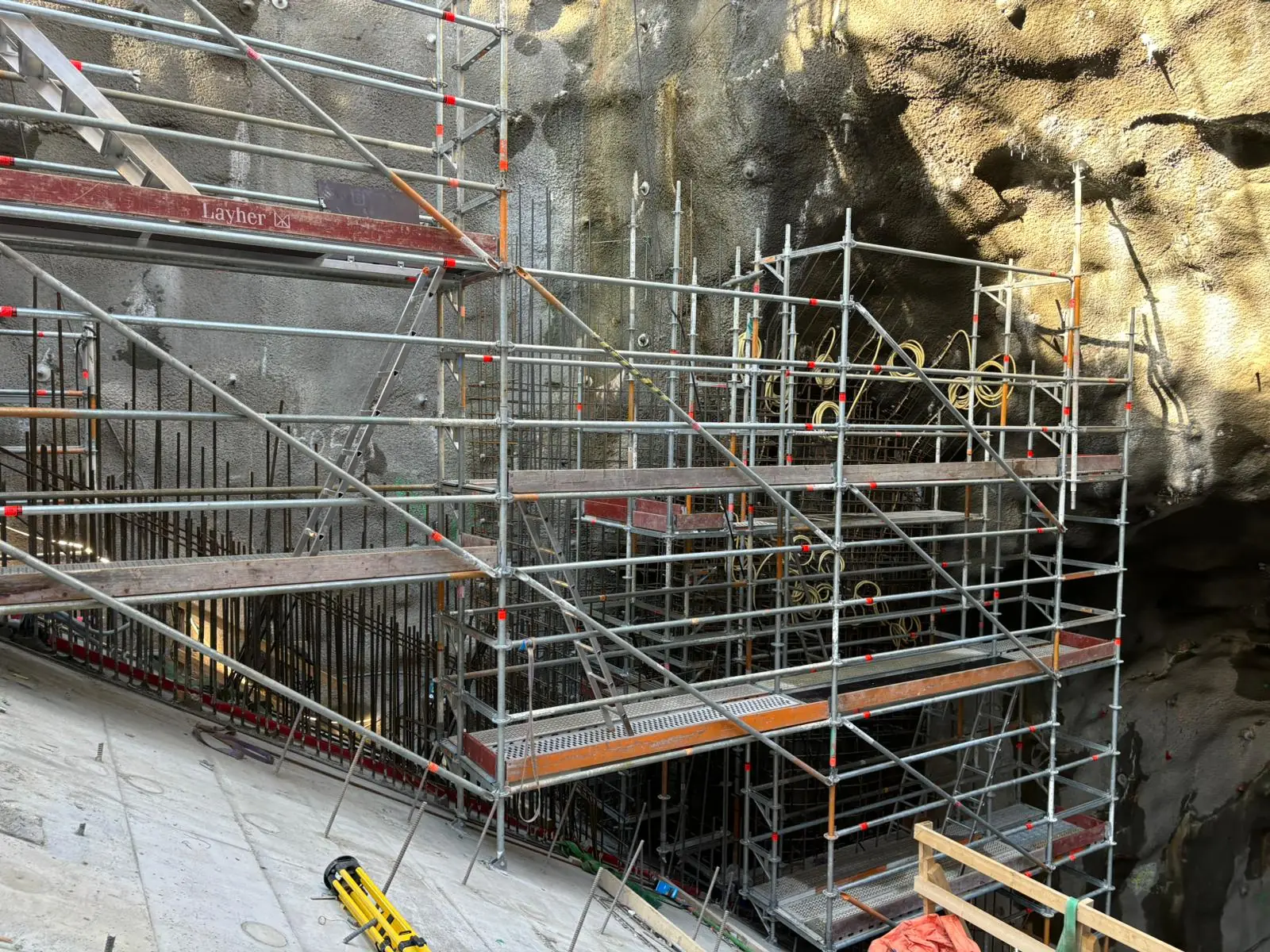 Ställning med arbetsplan mot en sprutbetongklädd bergvägg, med armering bakom och en tunnelöppning till höger